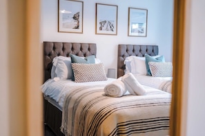 A hotel room with two beds, each with a tufted headboard, white bedding, and decorative pillows. There are framed pictures on the wall above the beds. - Centrally Located Flat off Formby High Street (Liverpool)