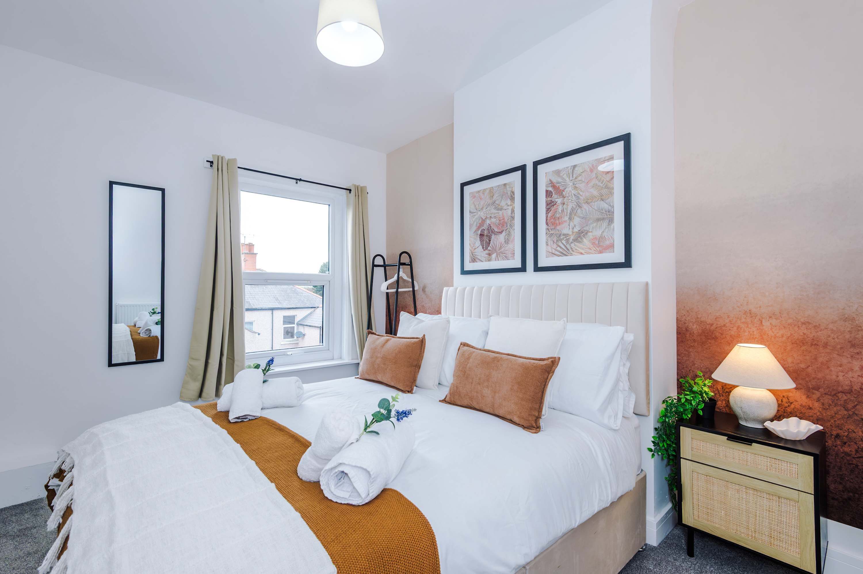 A hotel room with a bed, a bedside table with a lamp, a window with curtains, and framed artwork on the wall.