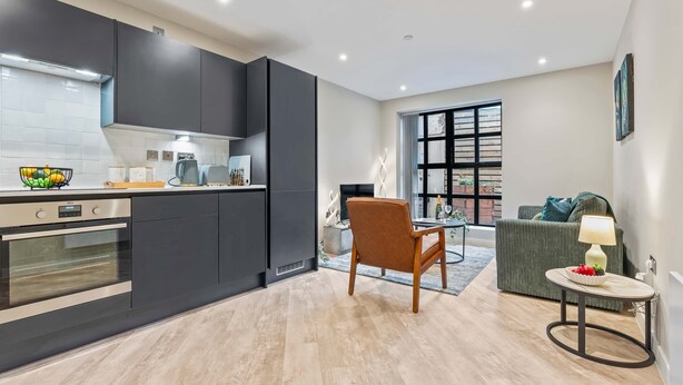 A modern kitchen with dark cabinets, a built-in oven, and a dining area with a table and chair. - Stylish 1 Bed Flat in Birmingham City Centre (Birmingham)