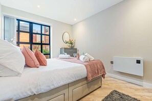 Ein Schlafzimmer mit Bett, Fenster, Spiegel, Heizkörper und einem Nachttisch mit Blumen.'
