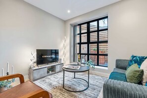 Ein modernes Wohnzimmer mit Fernseher, einem kleinen runden Tisch, einer Couch und einem großen Fenster.