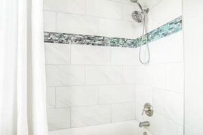 Un baño con azulejos blancos, una ducha con cabezal de ducha manual y un diseño de azulejos en esquina.