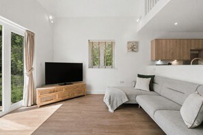 Ein modernes Wohnzimmer mit einem Flachbildfernseher auf einer Holzkonsole, einem grauen Sofa und einem großen Fenster mit Vorhängen.