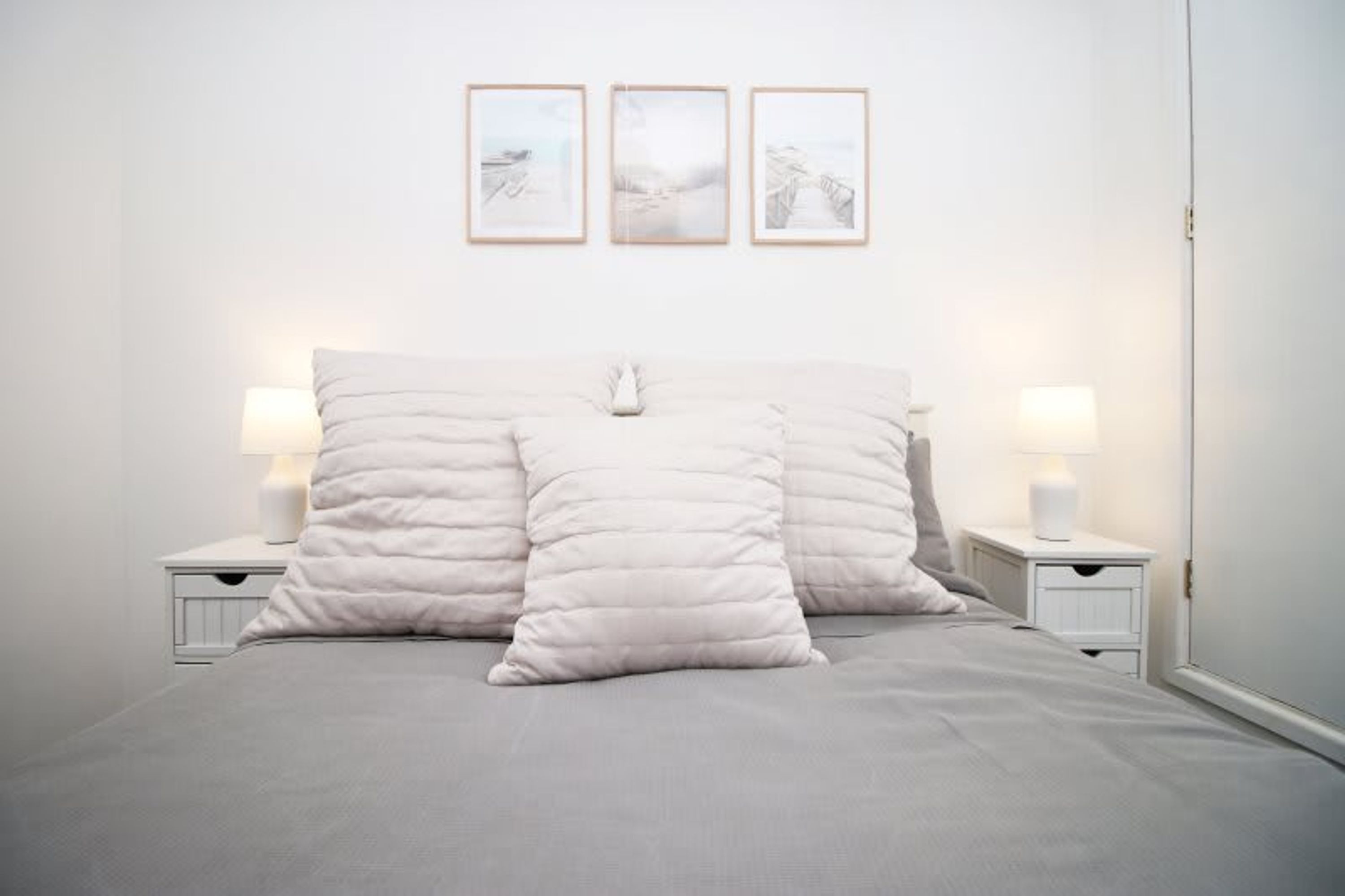 Cama bem arrumada com duas almofadas e duas luminárias de cabeceira, parede branca com três quadros e uma porta branca.