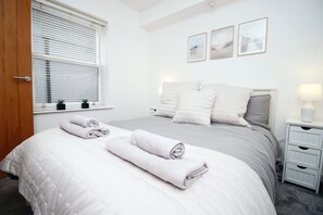 A bedroom with a bed, pillows, a bedside table, a lamp, a window with blinds, and three framed pictures on the wall. - Stylish 1-bed Flat at Irvine Court Sleeps 4 (Swansea)