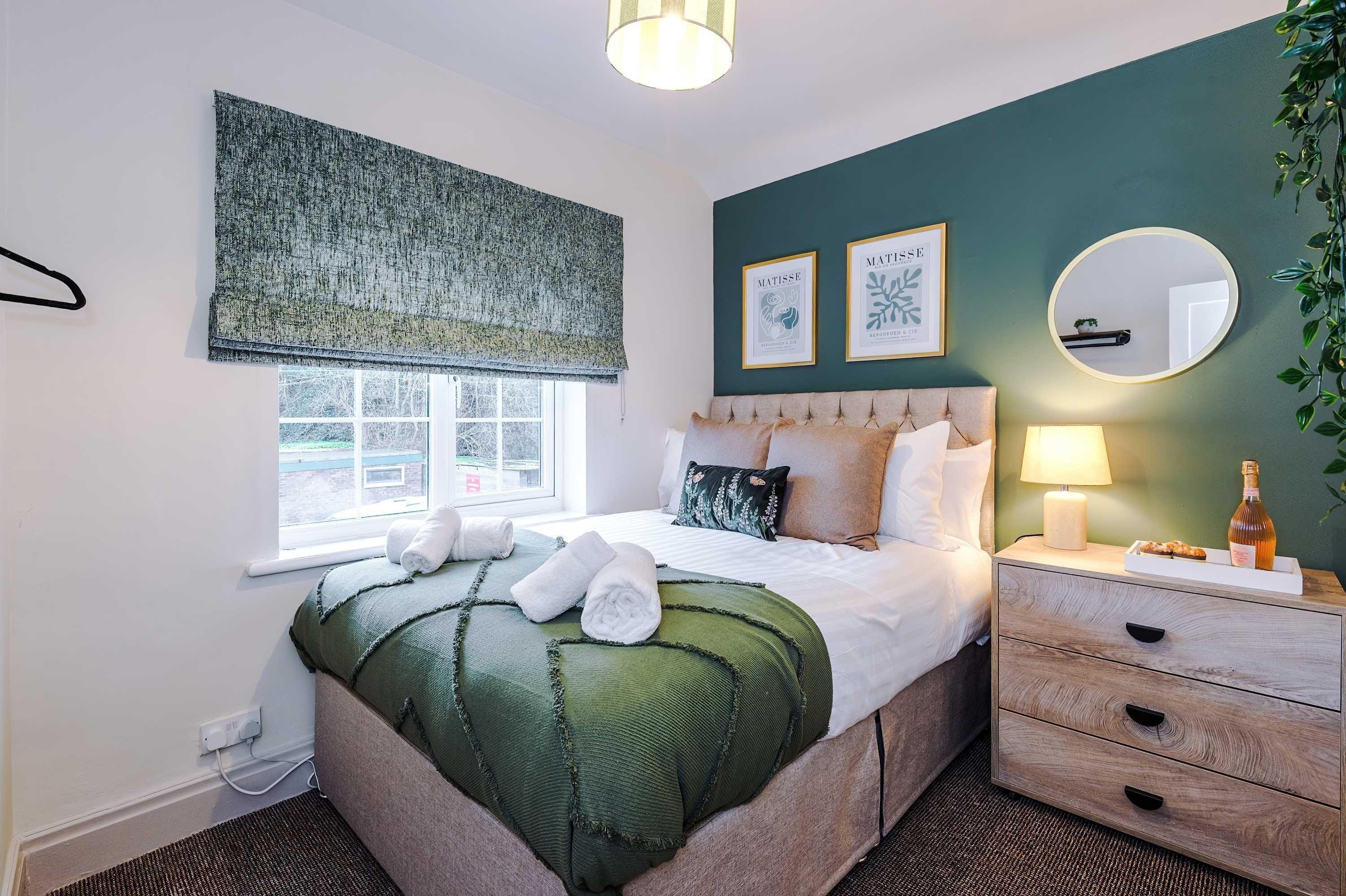 A neatly made bed with a green blanket, a bedside lamp, and a bottle on the nightstand.