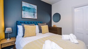 A bedroom with a blue upholstered headboard, a bed with white and patterned bedding, a bedside table with a lamp, and a wall-mounted mirror. - Charming City Centre 3 Bed Flat High St Location (Chester)