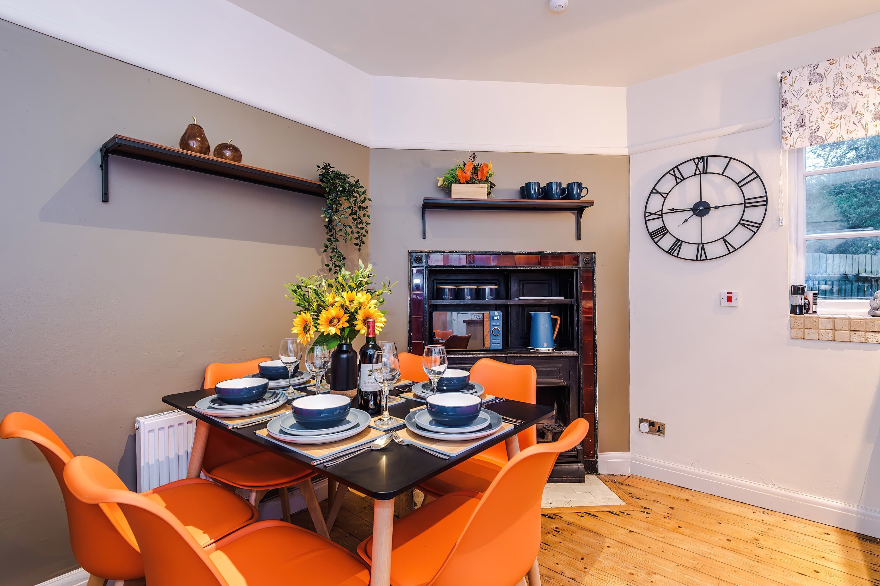 Un comedor con una mesa preparada para cuatro, sillas naranjas, mantel negro y un reloj en la pared.