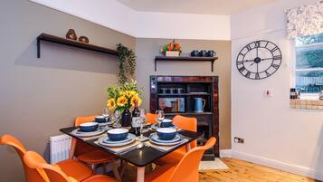 Un comedor con una mesa preparada para cuatro, sillas naranjas, mantel negro y un reloj en la pared.