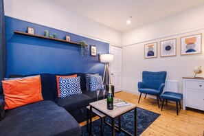 A modern living room with a blue accent wall, a sofa with patterned cushions, a small table with a bottle and glasses, a blue armchair, and a white cabinet. - Charming City Centre 3 Bed Flat High St Location (Chester)