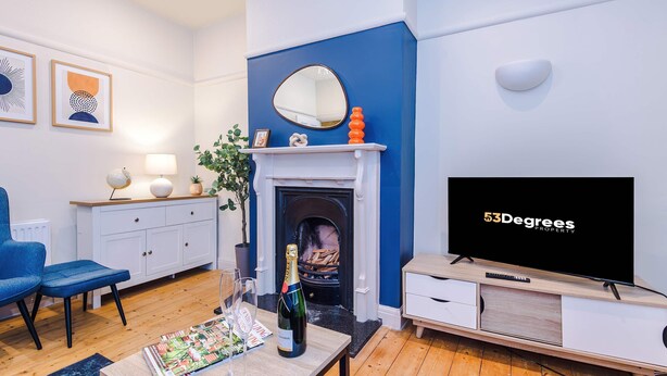 A living room with a fireplace, a television, a blue chair, a coffee table with a magazine and a bottle of champagne, and a white cabinet. - Charming City Centre 3 Bed Flat High St Location (Chester)