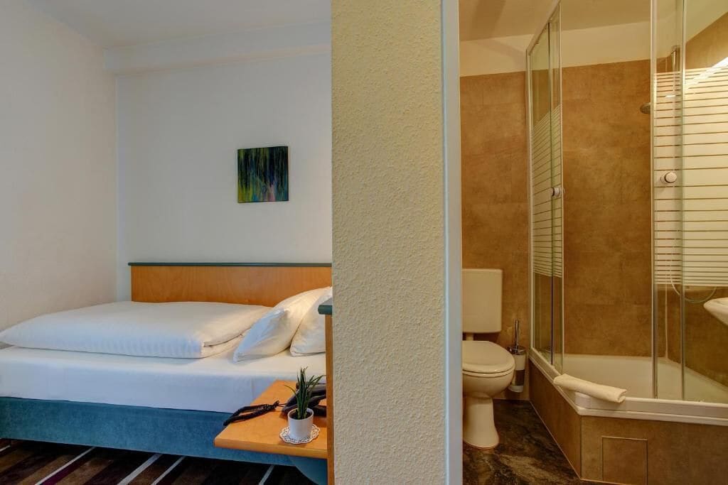 A hotel room with a bed, a small table with a plant, a toilet, and a glass-enclosed shower.