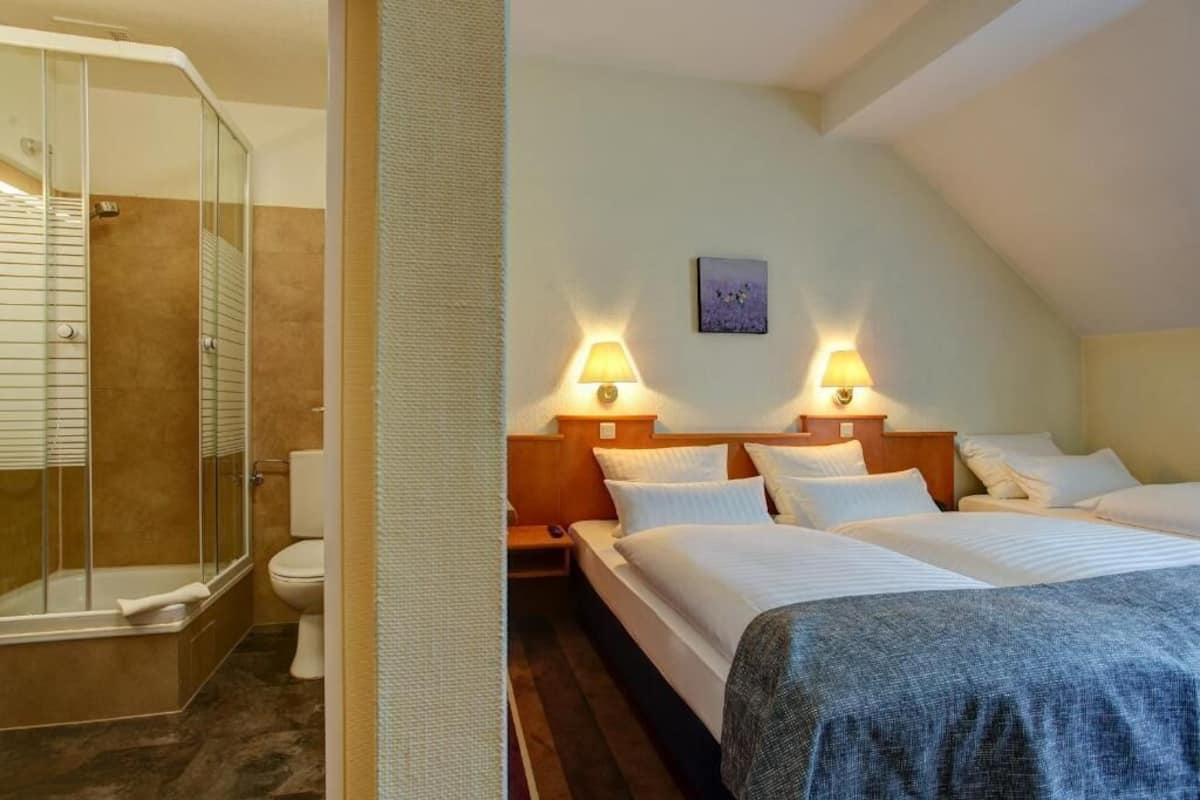 A hotel room with a shower, a bed, a bedside table, and a wall-mounted lamp.