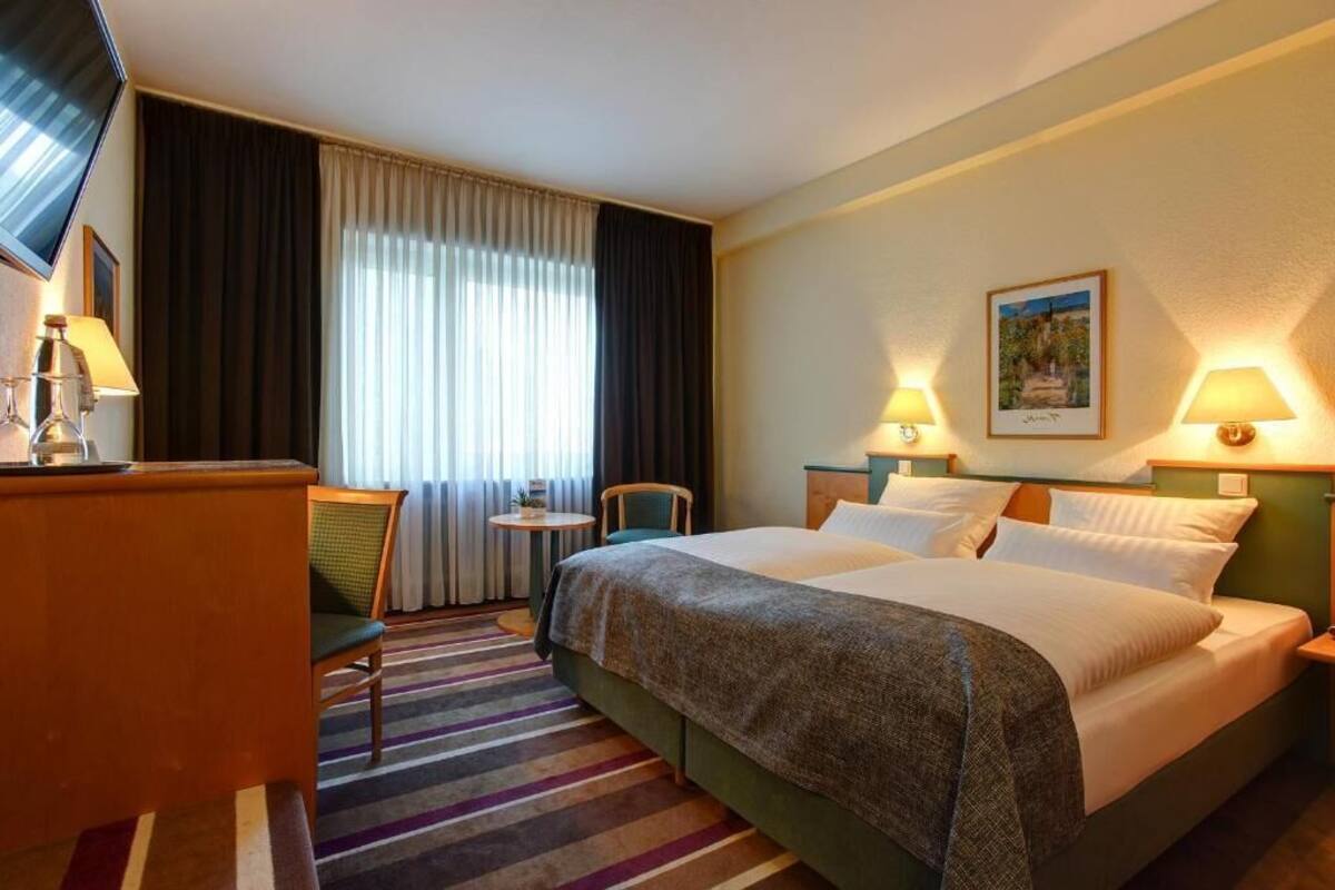A hotel room with a bed, a desk, a chair, a small table, and two wall-mounted lamps.