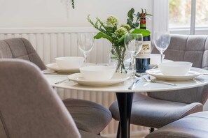 A dining table set with white plates, bowls, and wine glasses, accompanied by a bottle of wine and a vase with flowers. - Beautiful Large Home in Cradley Heath Sleeps 8 (Cradley Heath)