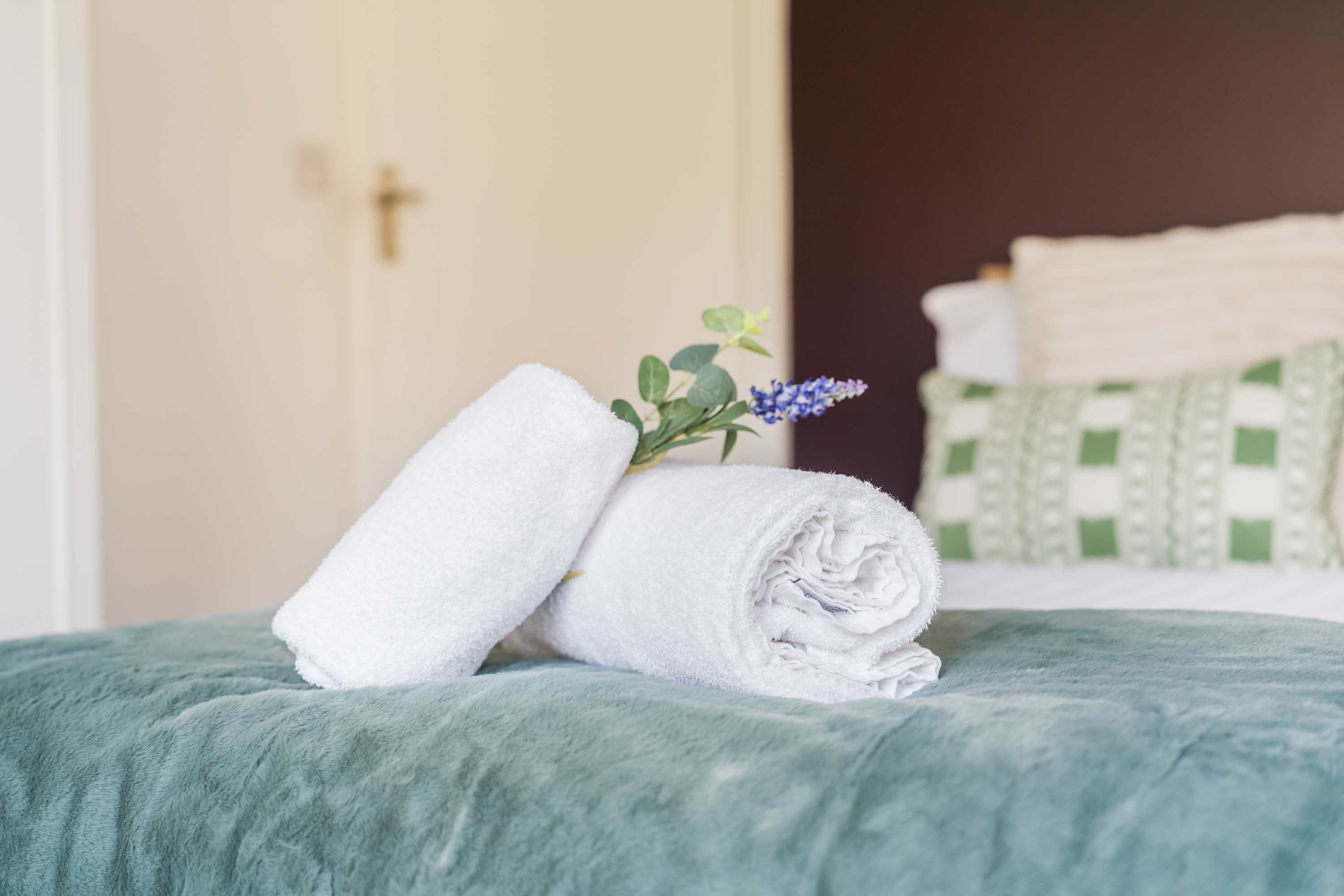 A neatly made bed with a green blanket, a rolled white towel, and a small plant with purple flowers.