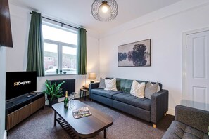 A modern living room with a flat-screen TV, a sofa, a coffee table, and a window with curtains. - Spacious 4 Bed Home in Crewe Perfect for Groups (Crewe)