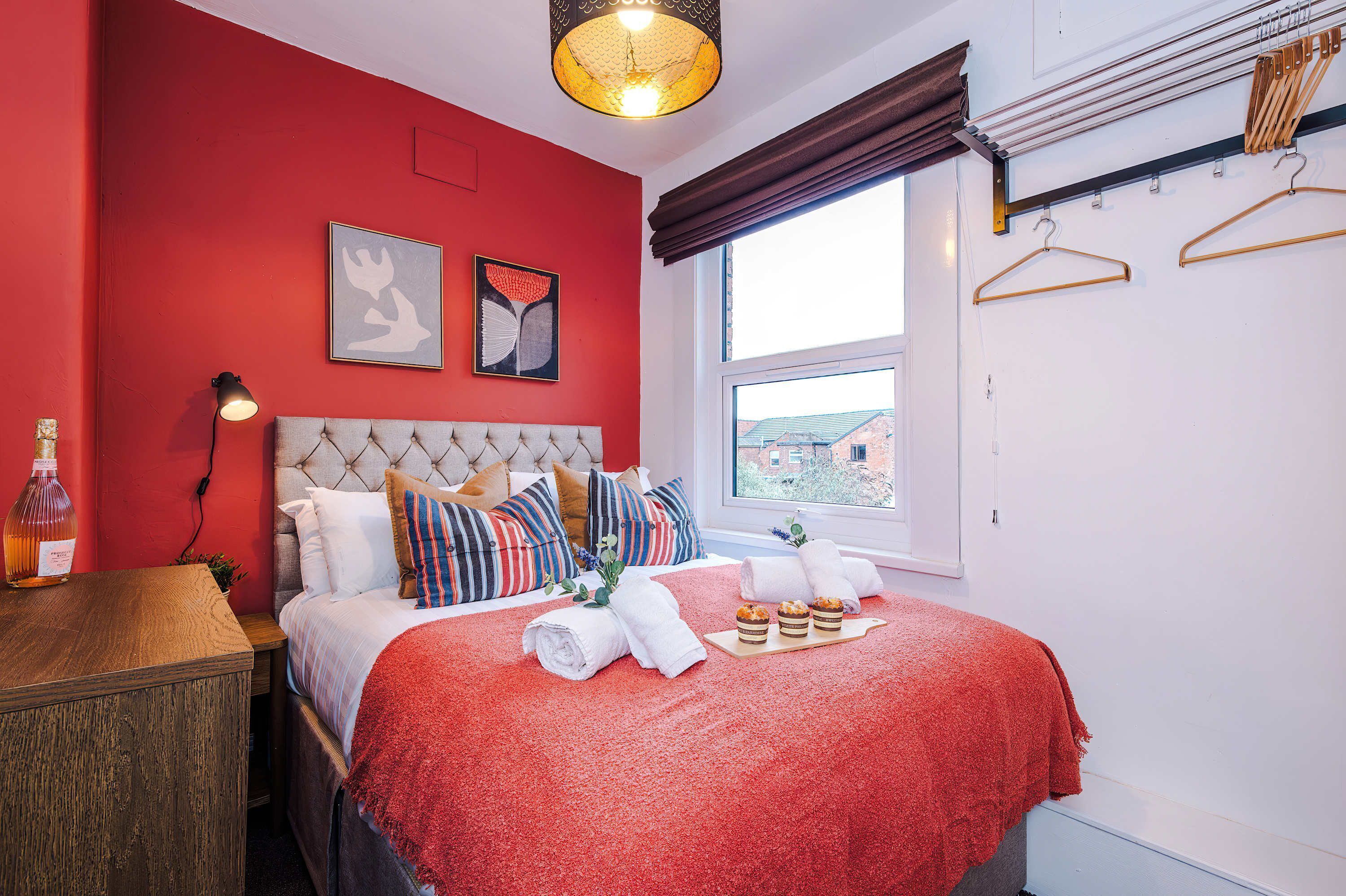 A bedroom with a red wall, a bed with a red blanket, a bedside table with a bottle, and a window with a view.