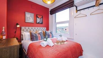 A bedroom with a red wall, a bed with a red blanket, a bedside table with a bottle, and a window with a view.
