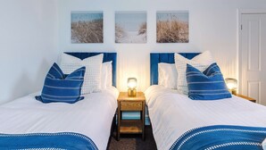 Una cama doble con ropa de cama blanca y detalles azules, dos mesitas de noche con lámparas y tres obras de arte con motivos playeros.