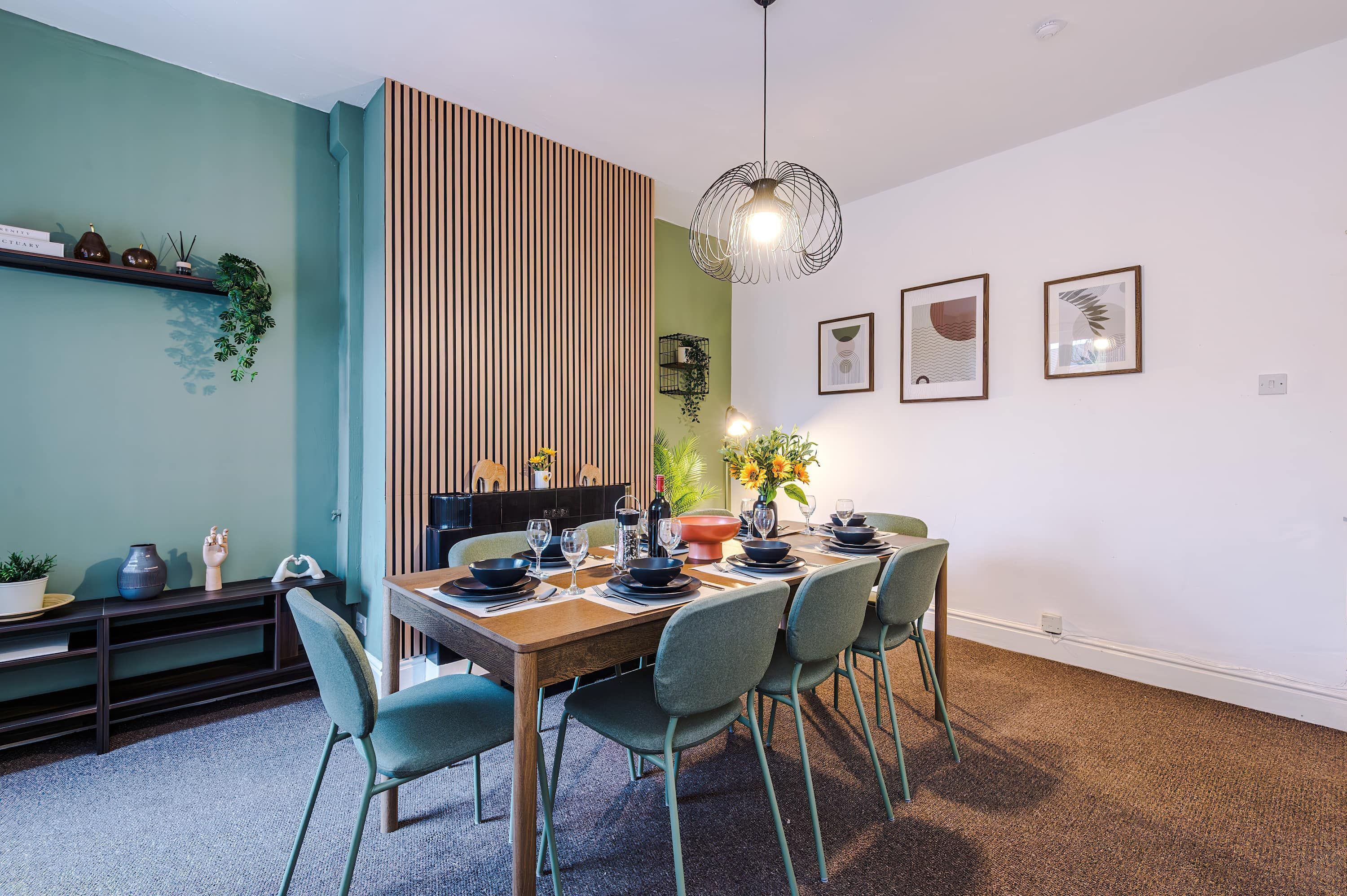A dining area with a wooden table set for four, surrounded by chairs, a hanging light fixture, and framed artwork on the wall.