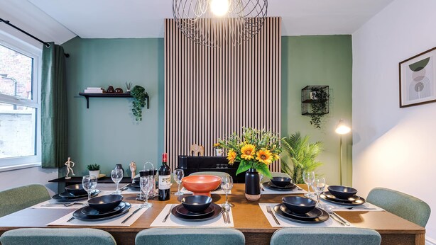 Comedor con mesa de madera dispuesta para cuatro personas, rodeada de sillas tapizadas en verde.
