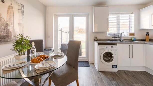 A modern kitchen with a glass dining table, a washing machine, and a view of the city through the window. - Modern 2 Bedroom Home in Swansea Near Stadium (Swansea)