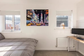 Ein Schlafzimmer mit Bett, Schreibtisch mit Computer, zwei Fenstern und einem Stadtbild.