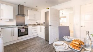 A modern kitchen with white cabinetry, stainless steel appliances, and a dining area with a glass table set for a meal. - Modern 2 Bedroom Home in Swansea Near Stadium (Swansea)