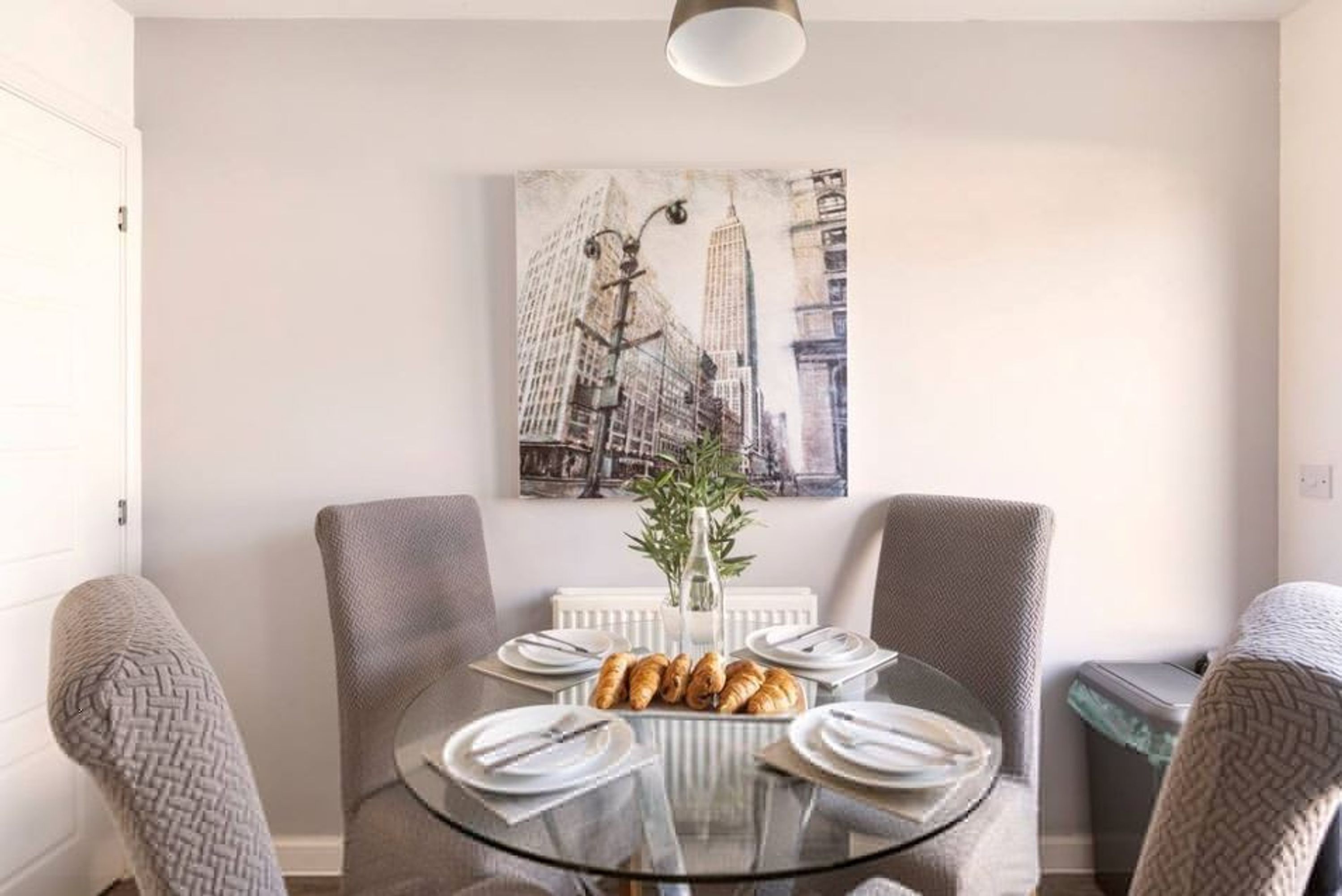 Un comedor con mesa de vidrio, sillas tapizadas grises, un radiador blanco y un cuadro con vista a la ciudad.