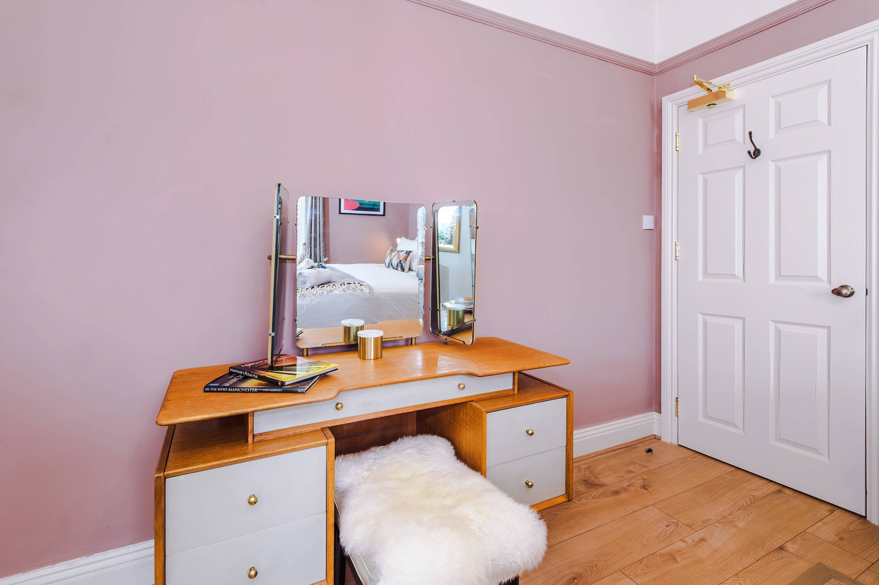 Una habitación con tocador de madera, un banco blanco y mullido, una puerta blanca y paredes rosadas.