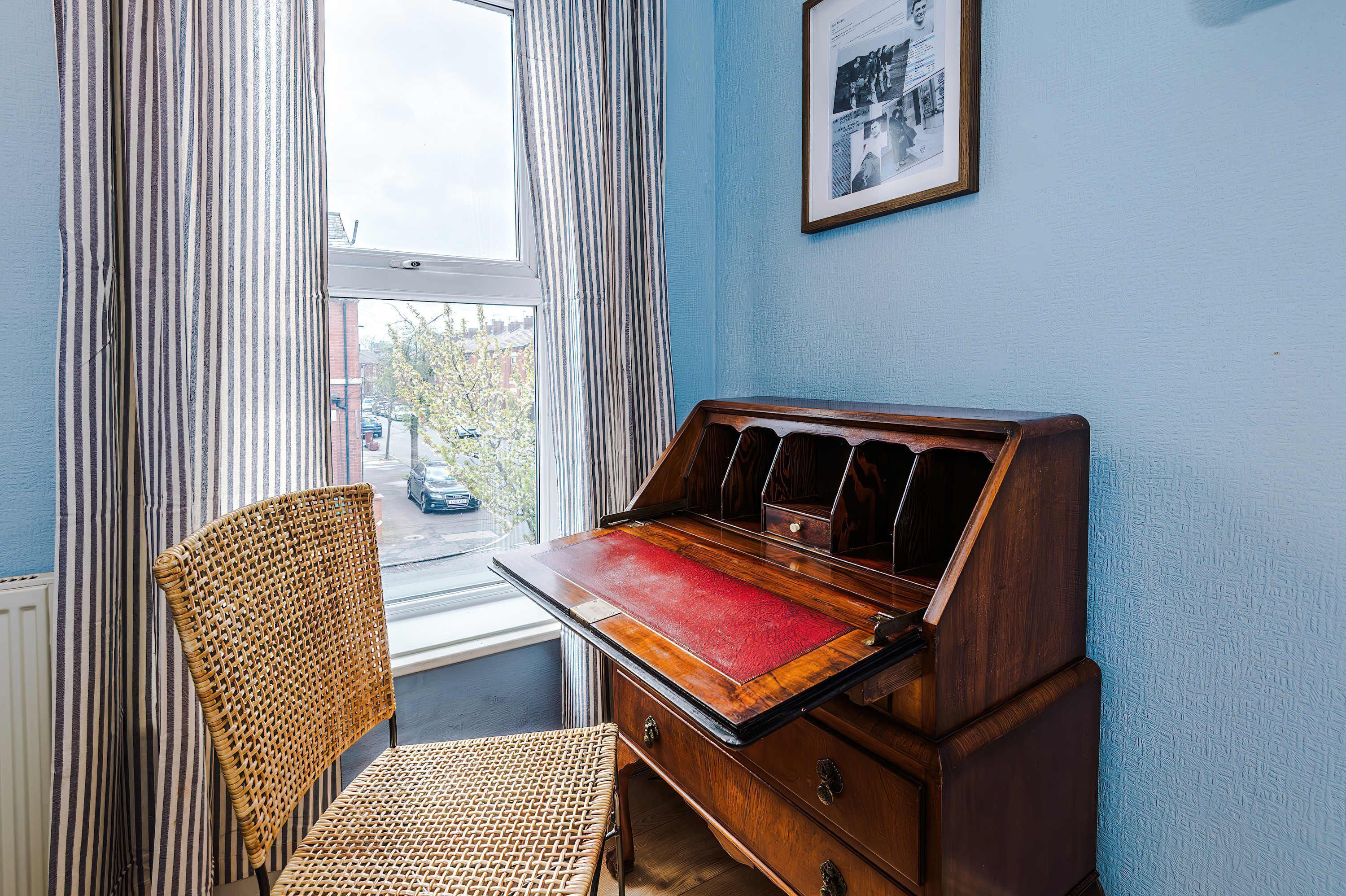 Een kamer met een houten bureau, een rieten stoel, een raam met gordijnen en een lijst met een afbeelding aan de muur.