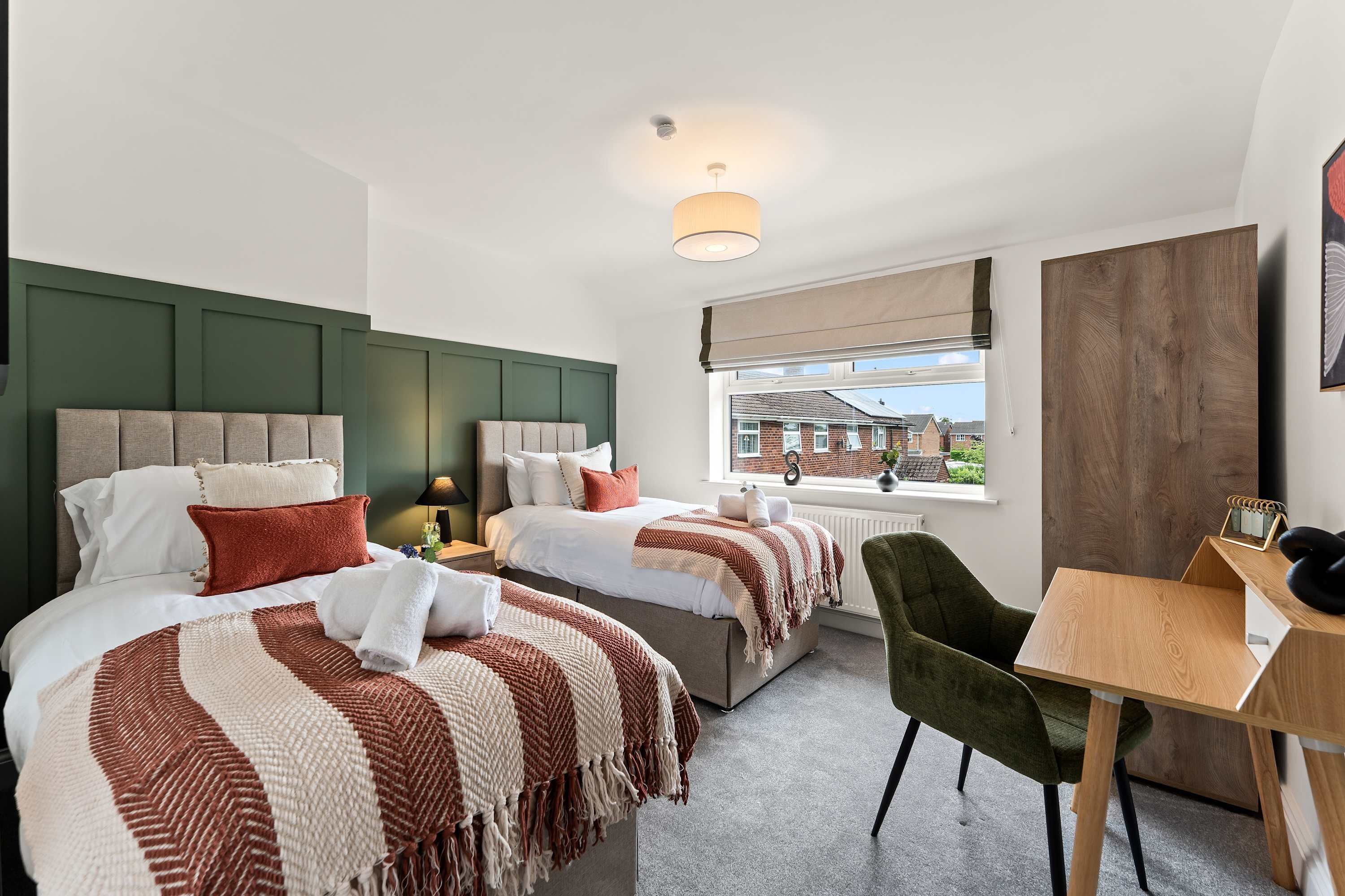 A hotel room with two beds, a desk, a chair, a window with a view, and a ceiling light.