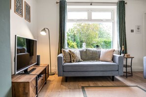 Una sala de estar con un sofá gris, un televisor sobre un mueble de madera, una lámpara de pie y una ventana con cortinas.