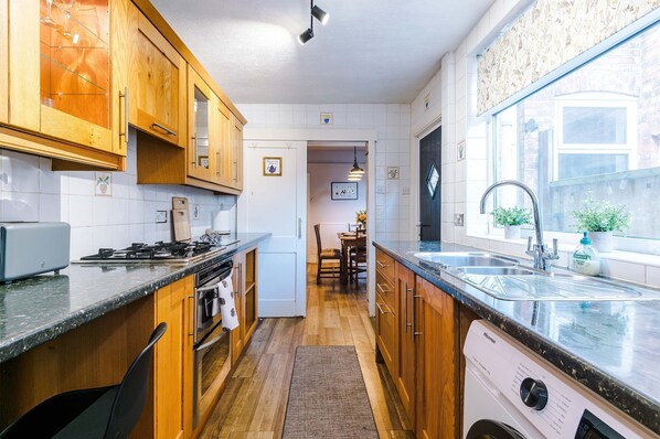 A kitchen with wooden cabinets, a built-in oven, a dishwasher, and a window with potted plants. - Spacious 3 Bed Perfect for Families and Groups (Nantwich)