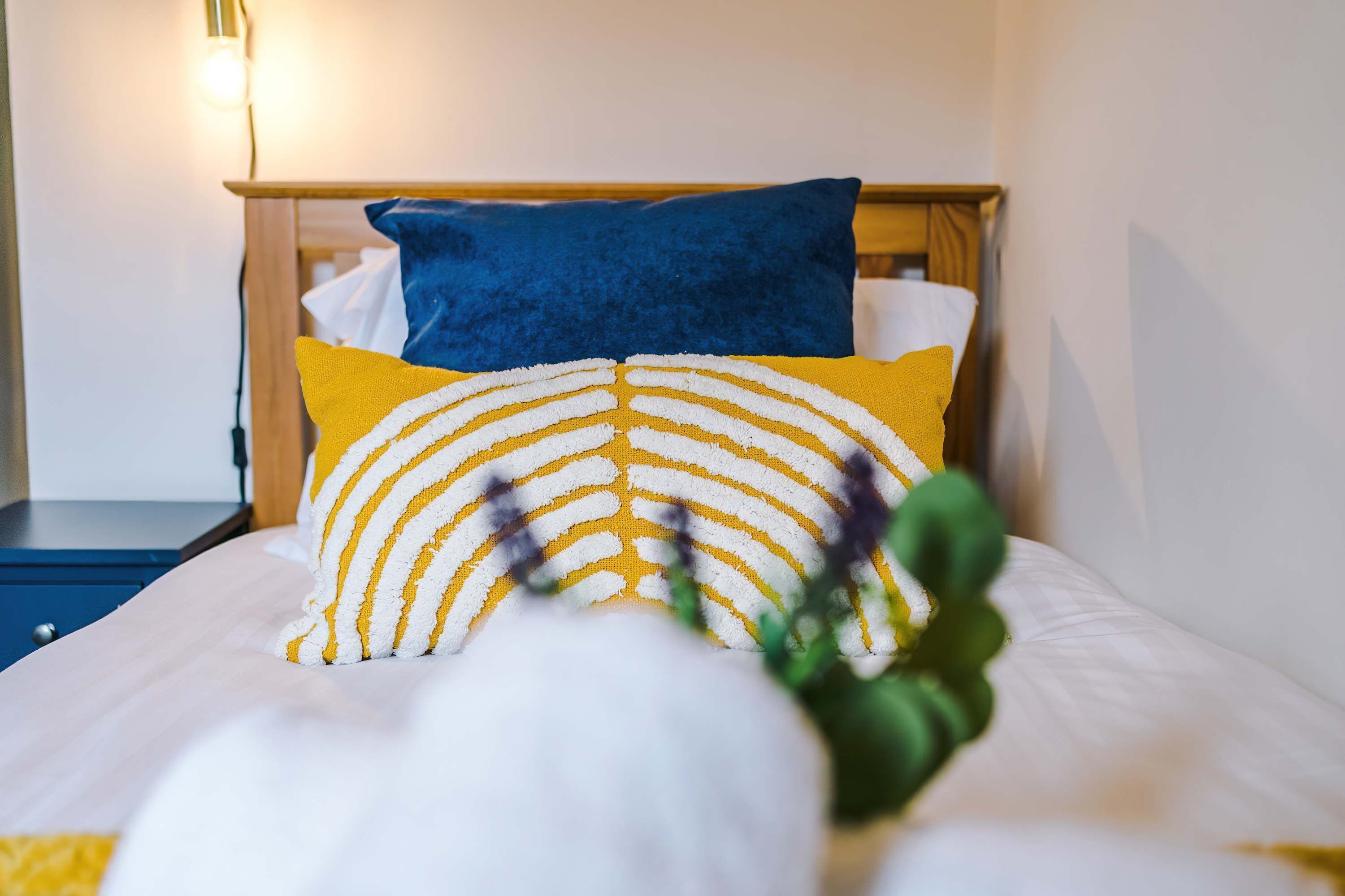 Una cama bien hecha con un cojín a rayas blanco y amarillo, un cojín azul y un cabecero de madera.