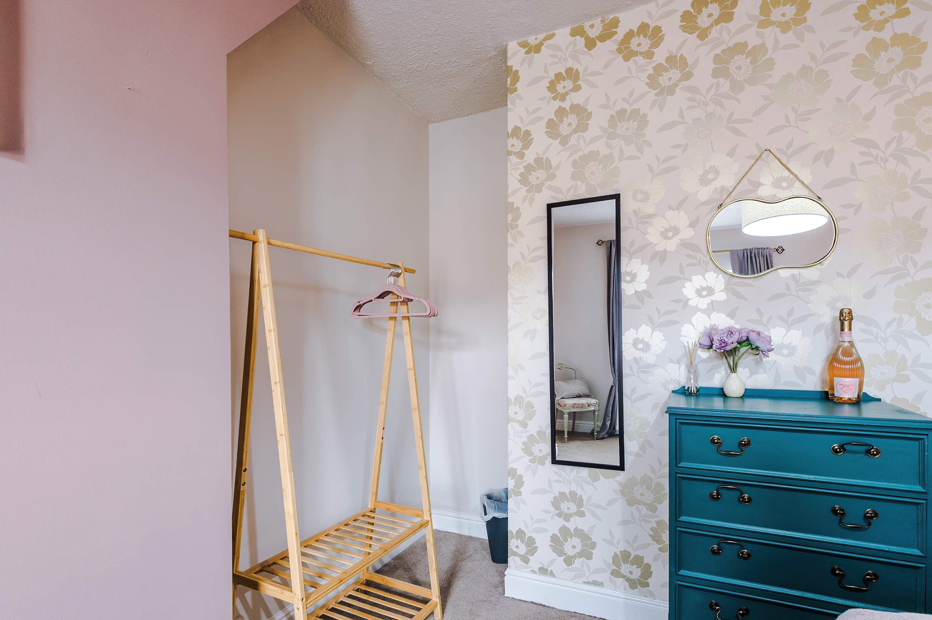 Une pièce avec un étendoir en bois, un miroir, une commode à tiroirs et un papier peint à motifs floraux.