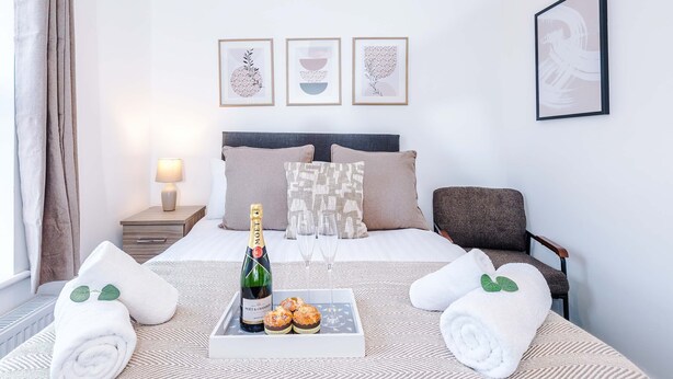 Quarto de hotel com cama, mesa de cabeceira com luminária, cadeira e bandeja com garrafa, copos e doces.