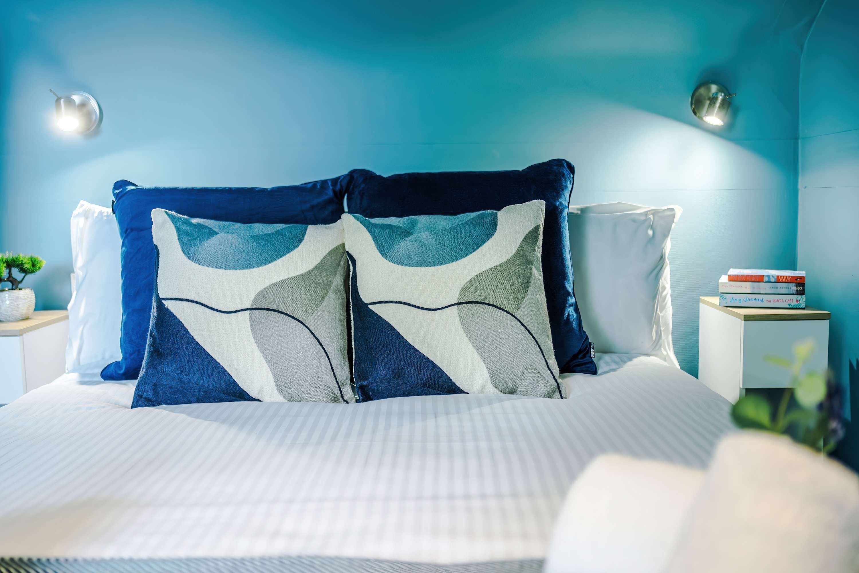 A bed with patterned pillows, a bedside table with books, and a wall-mounted lamp.