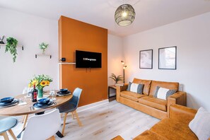 A modern living room with a wooden dining table, a sofa, and a TV mounted on an orange wall. - Perfect Modern 4 Bed in Crewe Great for Business (Crewe)