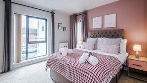Ein Hotelzimmer mit einem großen Bett, einem Nachttisch mit Lampe, einem Fenster mit Vorhängen und einem Bild an der Wand.