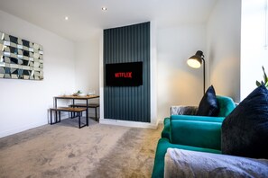 A living room with a flat-screen TV mounted on a wall, a small table with a plant, a green sofa, and a floor lamp. - Homely 2 Bed Close to Leeds City Center Sleeps 3 (Batley)