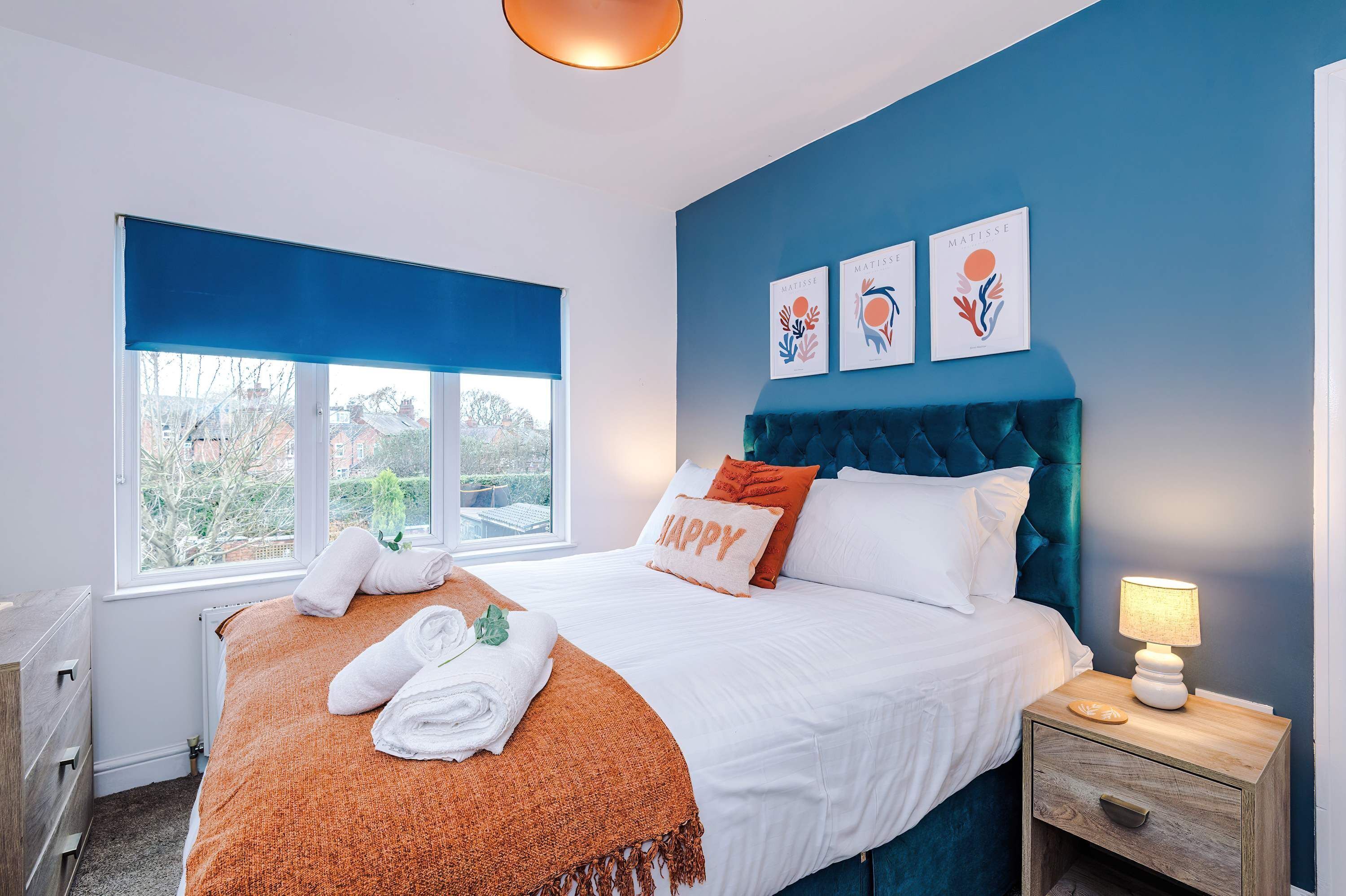 A bedroom with a bed, a bedside table with a lamp, a window with a view, and artwork on the wall.