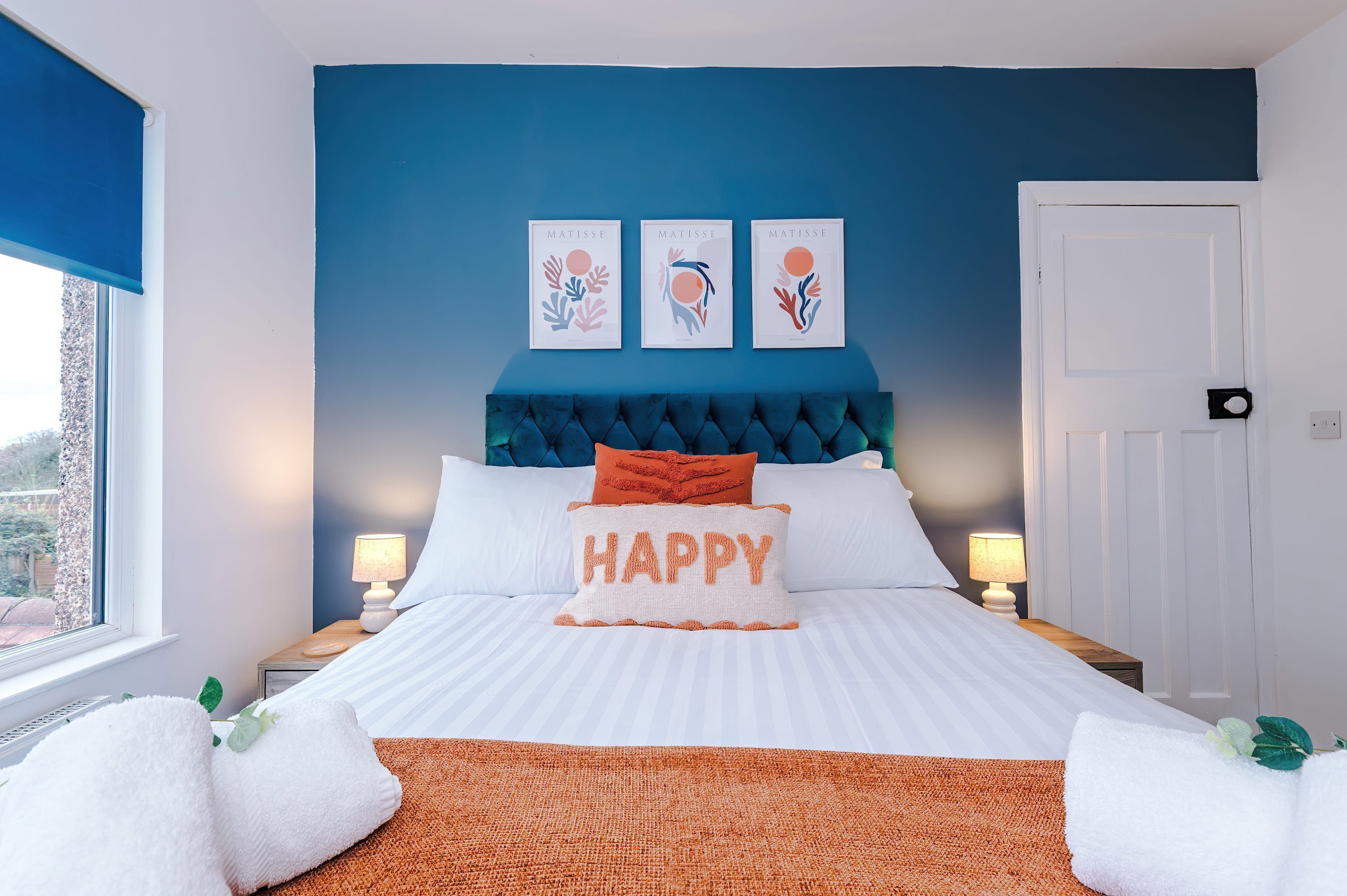 Un dormitorio con cama, pared azul, tres obras de arte enmarcadas, almohada con la palabra 'HAPPY' y dos lámparas de noche.