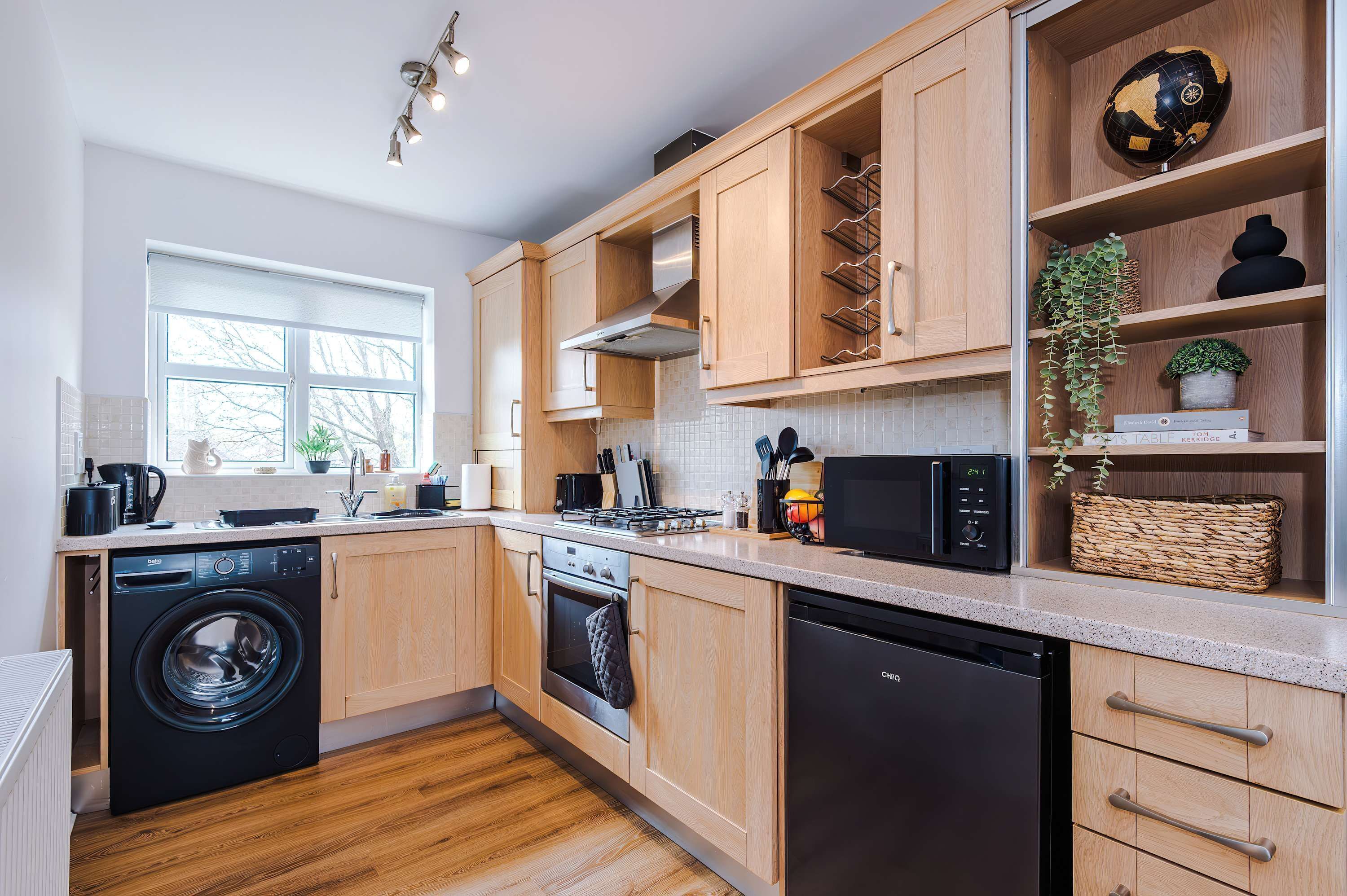 A modern kitchen with wooden cabinets, a dishwasher, a washing machine, and a window providing natural light.