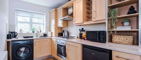 A modern kitchen with wooden cabinets, a dishwasher, a washing machine, and a window providing natural light.