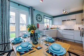 A dining area with a table set for four, a clock on the wall, and a kitchen in the background. - Spacious Modern 4 Bed in Northwich Free Parking (Northwich)