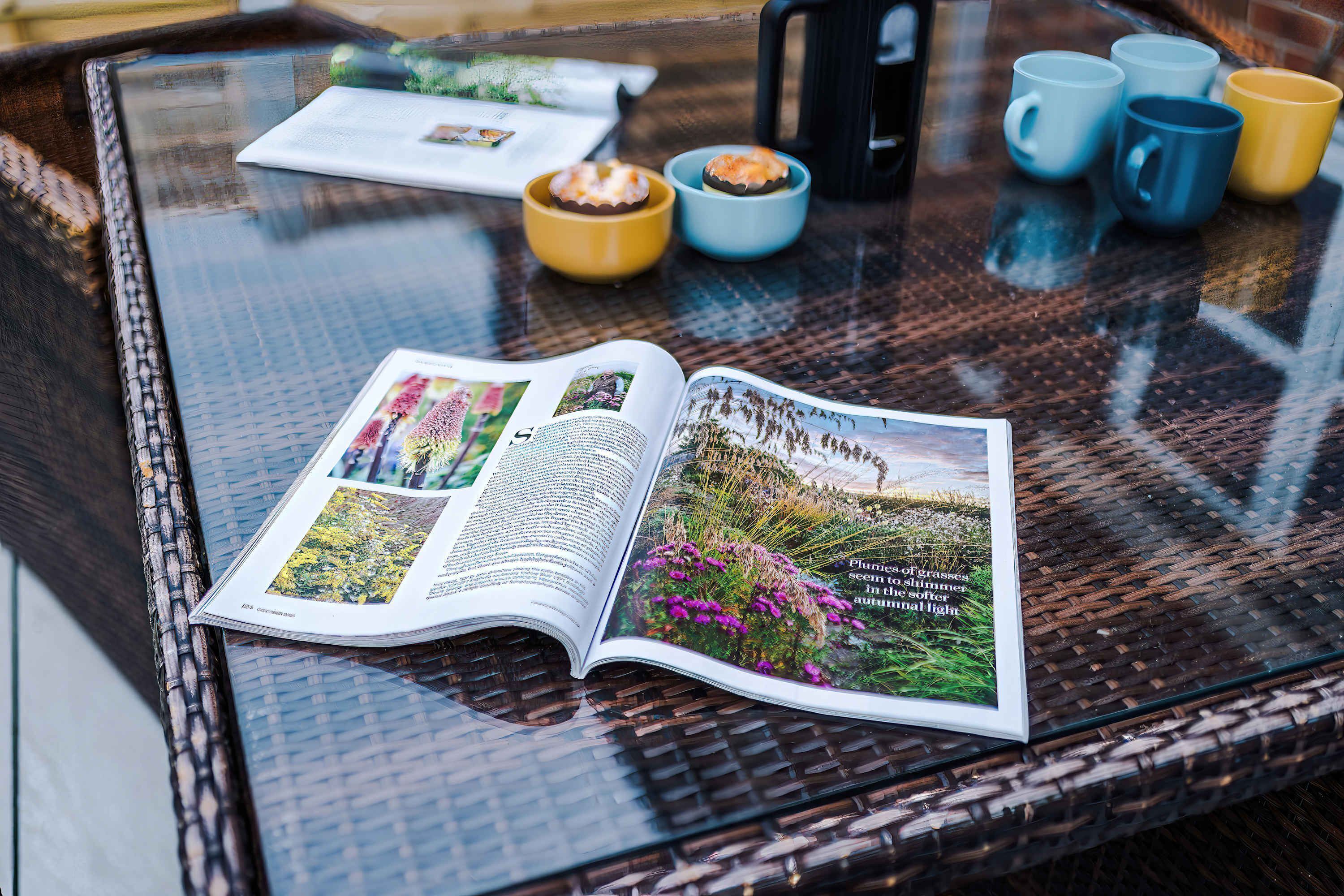 An open magazine on a glass table with a wicker chair, a yellow bowl, and several mugs.