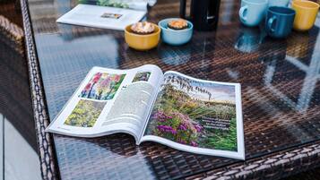 An open magazine on a glass table with a wicker chair, a yellow bowl, and several mugs.