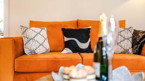 Ein Wohnzimmer mit einem orangefarbenen Sofa, gemusterten Kissen, einer Flasche und einer Auswahl an Gebäck.